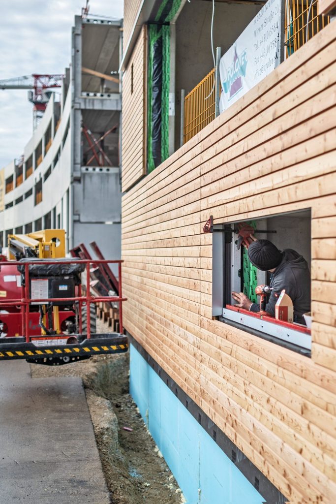 Montage der Holzfassadenelemente an den tragenden Stahlbetonfertigteilen der DOCKS im VILLAGE IM DRITTEN. Credit: ARE/Stefan Seelig